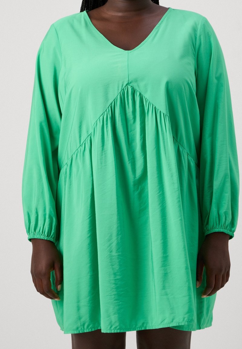 Grünes V-Ausschnitt-Kleid mit langen Ärmeln, geraffter Taille und fließendem Stoff. Die glatte Textur und das ausgestellte Design schaffen eine entspannte Silhouette.