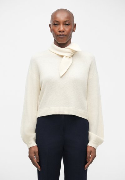 Cream knit sweater with a turtleneck and a tied scarf detail. Features wide sleeves and a cropped hem. Paired with dark trousers.