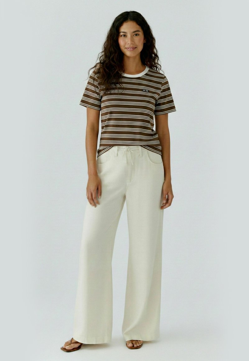 Femme aux longs cheveux bouclés portant un t-shirt rayé marron et blanc, un pantalon large crème et des sandales marron, debout et souriante.