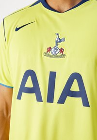 Neon gul Nike-trøje med mørkeblå accenter, som har et stort "AIA" logo og et klubmærke, der viser en hane over en bold.