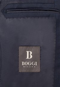 Jūras zila auduma ar gludu tekstūru, uz kura ir izšūta pelēka etiķete. Etiķete ir ar logo "B" un tekstu "BOGGI MILANO."