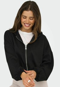Sweat à capuche noir zippé avec une coupe décontractée, présentant une capuche, des poignets côtelés et une texture lisse. Porté sur une chemise blanche.