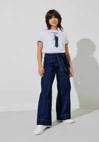 Hvid grafisk t-shirt med teksten "ICONIC", parret med højtaljede mørke denim bukser med vid ben og bælte i taljen. Hvide sneakers fuldender outfittet.