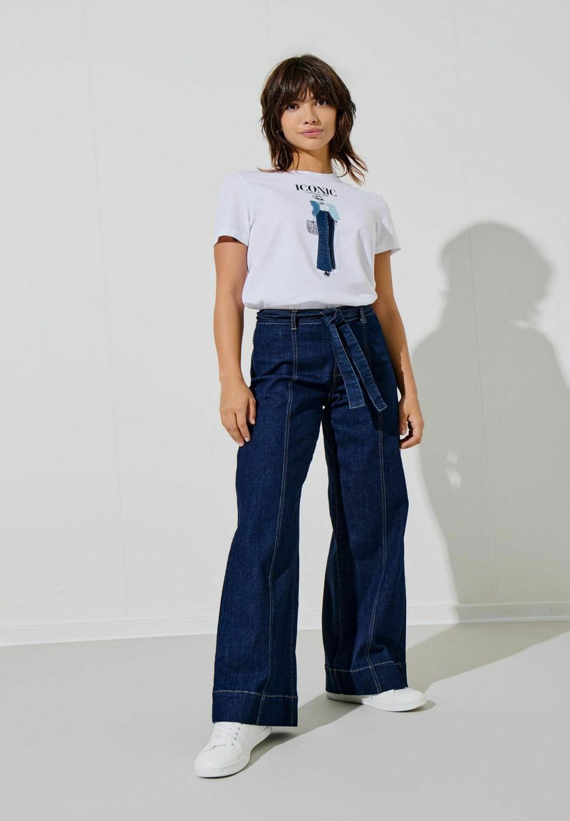 Hvid grafisk t-shirt med teksten "ICONIC", parret med højtaljede mørke denim bukser med vid ben og bælte i taljen. Hvide sneakers fuldender outfittet.