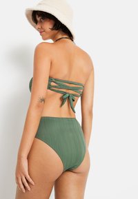 Žalias dviejų dalių bikinis su raukšlėta tekstūra. Viršus turi be rankovių dizainą su užrišamu uždarymu; aukšto liemens apatiniai užtikrina pilną uždengimą.