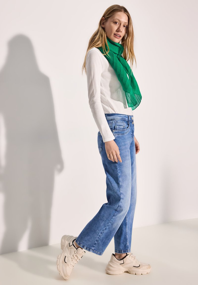 Blå denimjeans i kombination med en vit långärmad skjorta och en genomskinlig grön scarf. Skorna är beige sneakers med svarta detaljer.