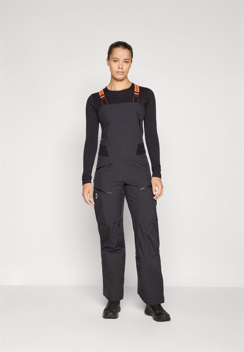 Schwarze Latzhose mit orangefarbenen Trägern, ausgestattet mit Reißverschlusstaschen und strukturierten Einsätzen. Kombiniert mit einem schwarzen Langarmshirt.