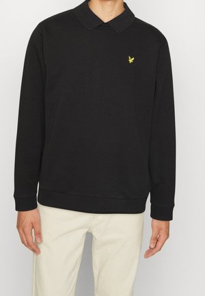 Mand iført sort langærmet polo med gul fuglelogo på brystet og lyse beige bukser, stående mod en ensfarvet baggrund.