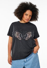 Camiseta de algodón negra con un gráfico de alas coloridas y texto en el centro. Mangas cortas y corte relajado. Combinada con pantalones cortos de mezclilla claros.