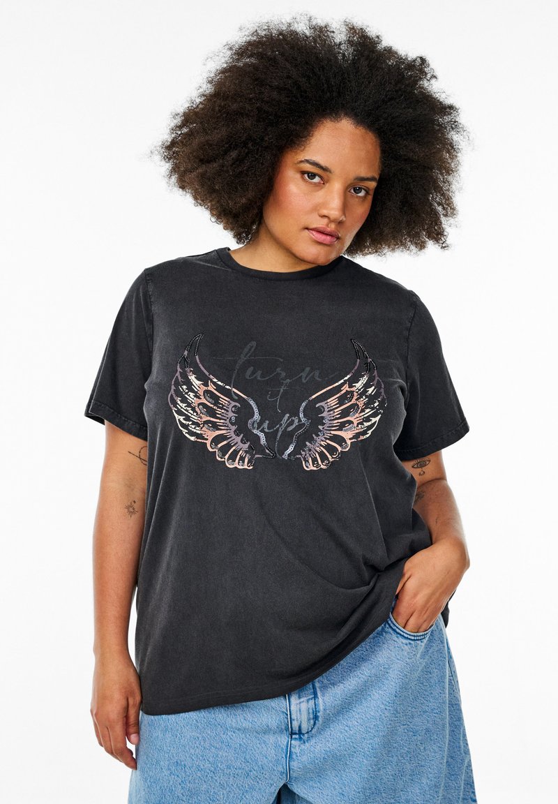 Camiseta de algodón negra con un gráfico de alas coloridas y texto en el centro. Mangas cortas y corte relajado. Combinada con pantalones cortos de mezclilla claros.