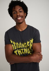 Musta puuvillainen t-paita, jossa on keltainen liekkityylinen "Stranger Things"-teksti. Lyhyet hihat, pyöreä kaula-aukko. Sileä pinta, istuva malli.
