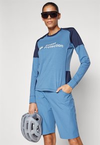 Zila garo piedurkņu krekls ar tumši zilu akcentu un logo, apvienots ar atbilstošām zilām šortiem. Modelis tur pelēku ķiveri. Gluda, viegla auduma.