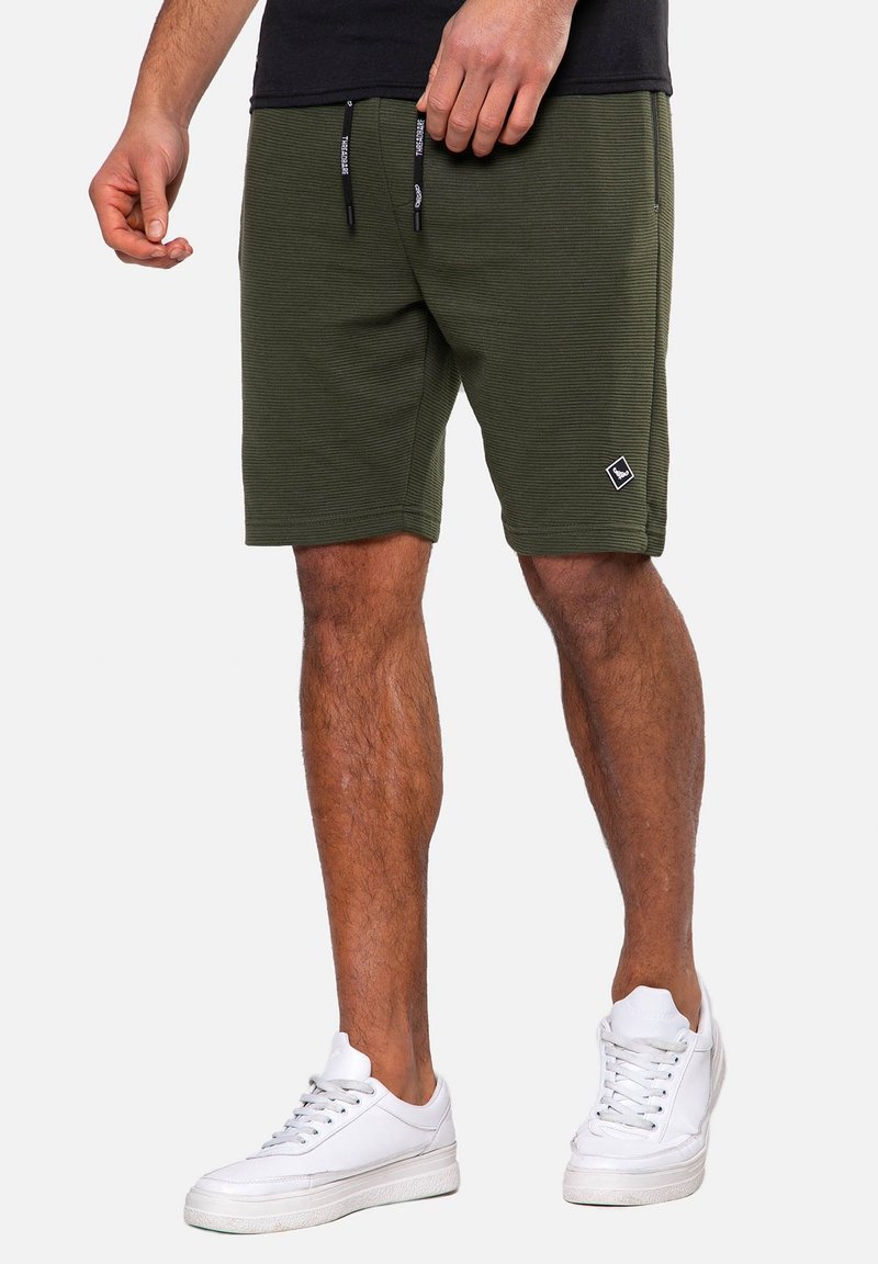 Shorts vert olive en tissu texturé, avec une taille élastique et des cordons de serrage, ornés d'un petit patch logo noir en bas.