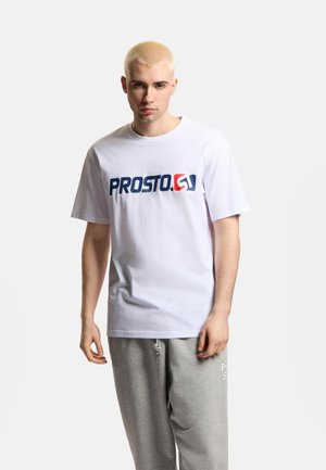 Młody mężczyzna z krótkimi blond włosami, ubrany w białą koszulkę z logo PROSTO i szare spodnie dresowe, stoi na jednolitym tle.