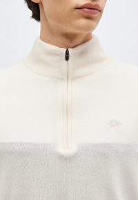 Muž oblečený v krémovom pulóvri s vysokým rebrovaným golierom a krátkym čiernym zipsom pri krku.