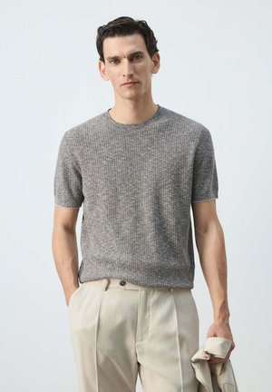 Hombre que lleva un suéter gris texturizado de manga corta y pantalones beige, sosteniendo una chaqueta beige en su mano derecha.