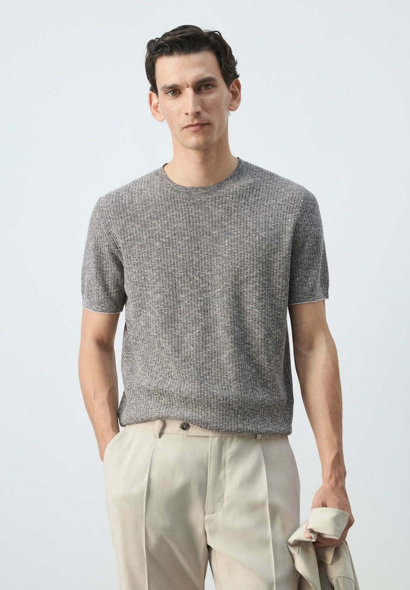 Homme portant un pull gris texturé à manches courtes et un pantalon beige, tenant une veste beige dans sa main droite.