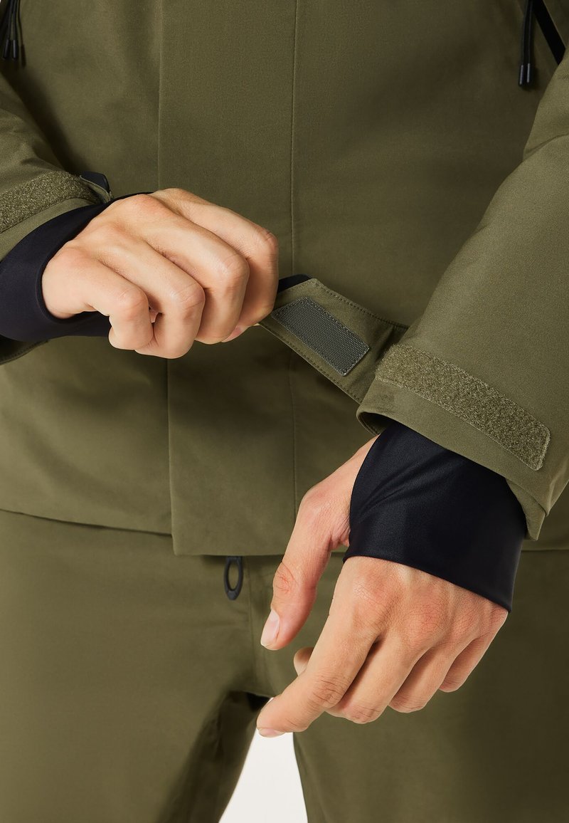 Olivgrüne Jacke mit Reißverschlusstasche und Klettverschluss. Schwarzes Innenfutter sichtbar an den Ärmeln. Hände, die die Ärmelbündchen anpassen.