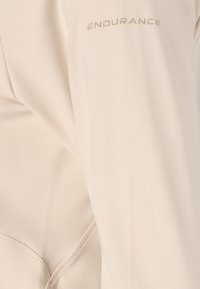 Helles beige Sportjacke mit glatter Textur, ausgestattet mit flachen Nähten und dem Wort "ENDURANCE" in einem helleren Farbton auf dem Ärmel gedruckt.