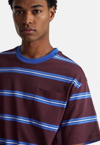Camiseta de manga corta burdeos con rayas horizontales azules y celestes, cuello redondo y un bolsillo en el pecho del lado izquierdo.