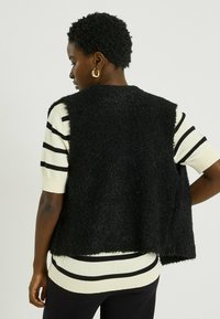 Chaleco negro y esponjoso con un ajuste holgado, superpuesto sobre una camisa a rayas crema. Presenta un dobladillo curvo y amplias sisas. Se ven pendientes de aro dorados.