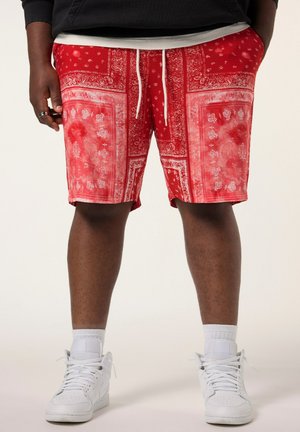 Person iført røde mønstrede shorts med snøre, hvide høje sneakers og hvide sokker, stående mod en ensfarvet baggrund.