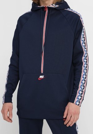 Persona vestida con sudadera Nike azul marino con cremallera roja y franjas blancas, azules y rojas con patrón en las mangas, de pie con las manos relajadas.