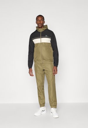 Veste zippée pour homme en noir, kaki et crème, avec une capuche et des blocs de couleur contrastants ; associée à un pantalon kaki assorti et des baskets blanches.