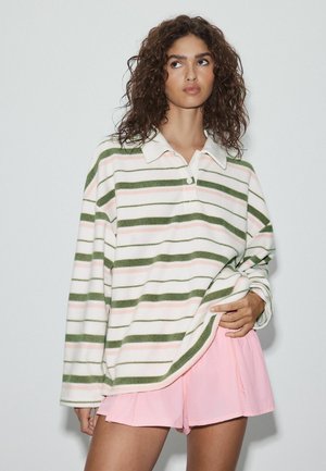 Vrouw met krullend haar die een crèmekleurig shirt draagt met groene en roze strepen en lichtroze korte broek, staand tegen een effen achtergrond.