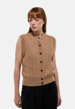 Femme aux cheveux roux lisses portant un gilet sans manches en tricot marron boutonné avec un pantalon noir, regardant vers le bas sur un fond uni.