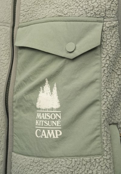 Zelená fleecová vesta s textúrovaným povrchom. Obsahuje predné vrecko, kruhový gombík a biele potlačené logo "MAISON KITSUNÉ CAMP."