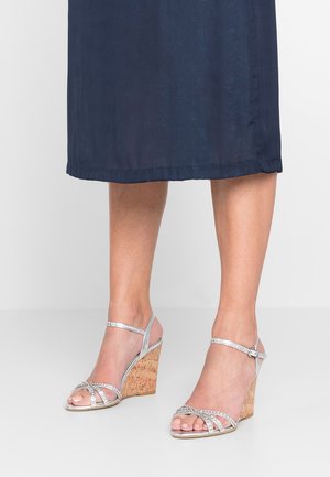 Srebrne sandały na koturnie z korkowym obcasem, krzyżującym się wzorem pasków oraz regulowanym zapięciem na kostce, z subtelnymi akcentami błyszczącymi.