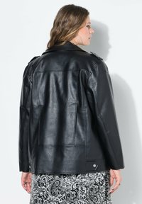 Chaqueta de imitación de piel negra con un diseño ajustado, acentos prominentes en los hombros y una textura sutil. La parte trasera cuenta con costuras horizontales y verticales.