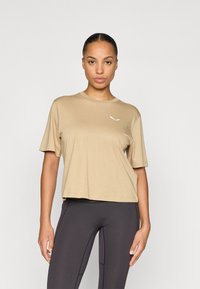 Camiseta de manga corta beige en tela suave, con detalle de logo blanco en el lado izquierdo del pecho y un ajuste holgado, ligeramente recortado.