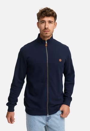 Hombre que lleva una chaqueta de cremallera en color azul marino con un pequeño parche de cuero en el pecho y jeans de color azul claro, de pie contra un fondo blanco.