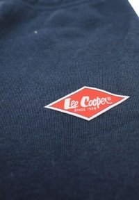 Tissu texturé bleu marine avec un patch blanc en forme de losange, présentant un logo rouge "Lee Cooper" et le texte "depuis 1908".