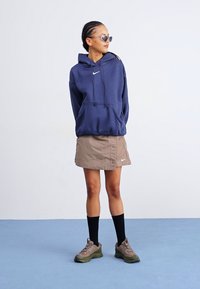 Sweatshirt azul-marinho com cordão e bolso frontal, saia bege com detalhes em botão, meias pretas até ao joelho, ténis cinza-esverdeados com solas grossas.