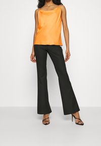 Orange satin linne med rundad halsringning, tillsammans med svarta utsvängda byxor. Modellen bär svarta sandaletter med remmar.