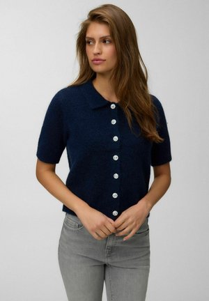 Frau mit langen braunen Haaren, die eine marineblaue kurzärmelige Strickjacke mit dekorativen Knöpfen und hellgraue Jeans trägt und zur Seite schaut.