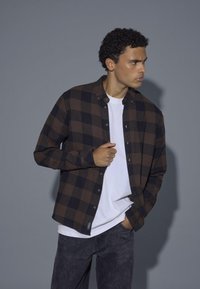 Rjava in črna karirana flanel srajca, zasnova z gumbi, nosi se čez belo majico. Sive kavbojke z diskretno teksturo. Model pozira s strani.