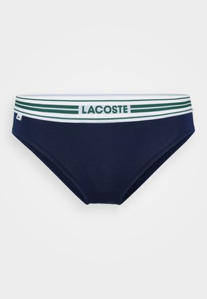 Mørkeblå svømmebukser med et elastisk liv bånd i hvide og grønne striber, der har "LACOSTE" logo. Glat tekstur og tætsiddende design.