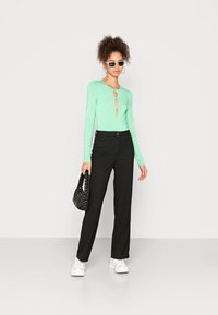 Camisola verde-menta de manga longa com aberturas em forma de olho-chave, combinada com calças largas pretas. O modelo segura uma bolsa trança preta e usa ténis brancos.