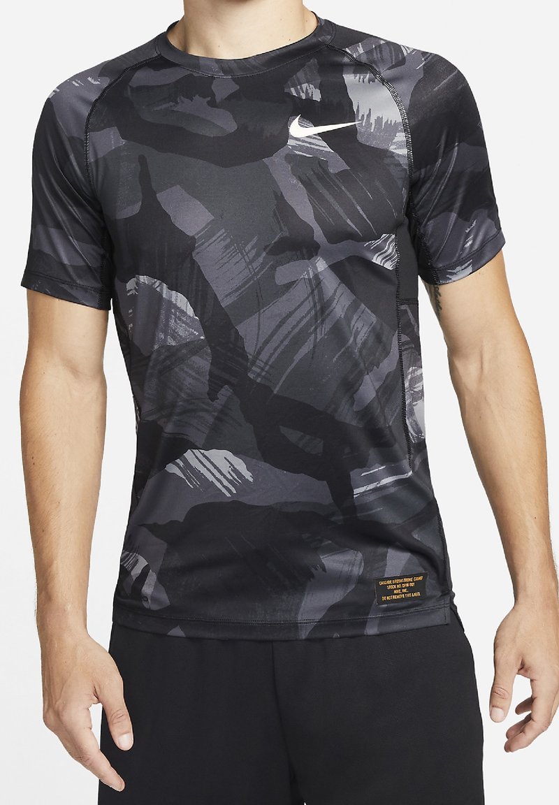 Homme portant un t-shirt de sport Nike à manches courtes avec un motif abstrait noir et gris et un short noir, debout les bras détendus.