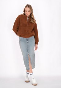 Hnědá fleecová bunda s kulatým límcem a předními knoflíky, spárovaná se světle modrou džínovou sukní s předním rozparkem. Model nosí bílé tenisky.