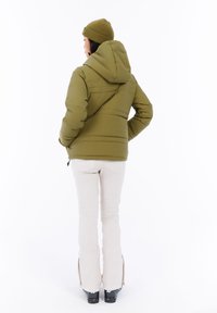 Vrouw in olijfgroene pufferjas, bijpassende beanie, witte broek en zwarte skies, met de rug naar ons gekeerd en handen in de zakken van de jas.