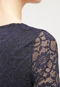 Gros plan sur l'épaule d'une personne portant un vêtement en dentelle florale bleu foncé avec des parties transparentes sur la manche et le haut du dos.
