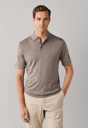 Young man standing with hand in pocket wearing taupe short-sleeve polo shirt and beige pants against a plain light background.
