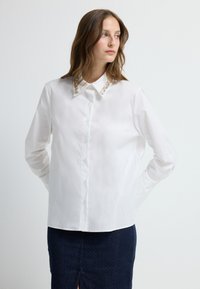 Witte blouse met kraag en heldere versieringen; lange mouwen, gladde stoftextuur, gecombineerd met een marineblauwe rok.
