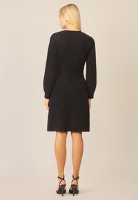 Sort strikket kjole med lange, puffede ærmer. Har et struktureret kabel-strikket mønster på bodice'en og en tætsiddende talje, der går over i en flagrende nederdel.