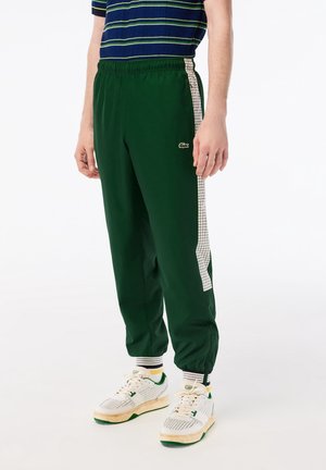 Homme portant un pantalon de survêtement vert foncé avec une bande latérale à carreaux blancs et des baskets blanches avec des touches vertes.